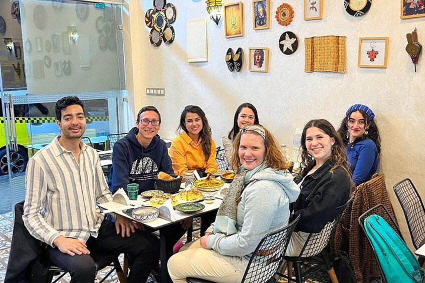 A group of people seated around a table with various dishes, in a room with decorative wall hangings. 1