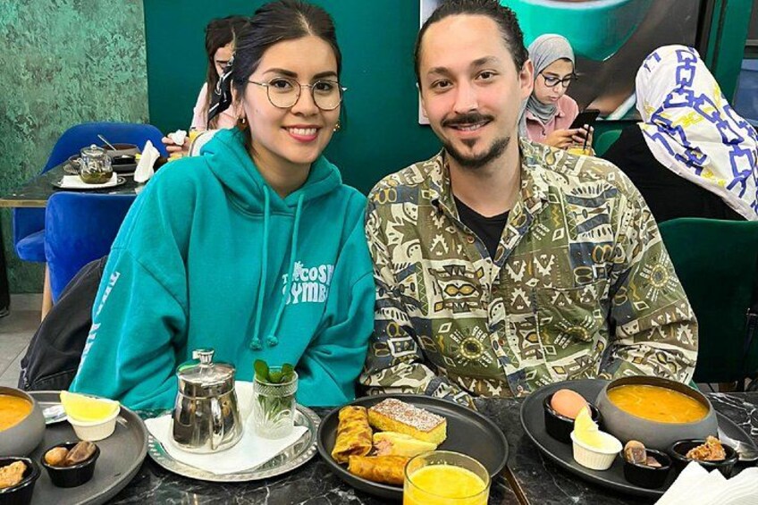 Two people seated at a table with various dishes, including a bowl of soup, a plate of food, and a glass of juice. 2