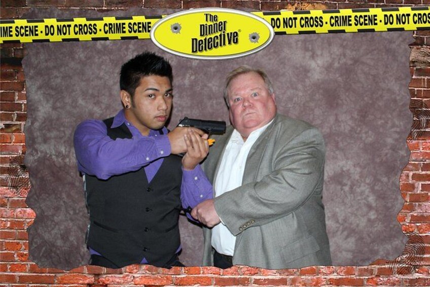 A man in a suit holding a toy gun, another man in a vest, and a backdrop with a crime scene tape border. 5