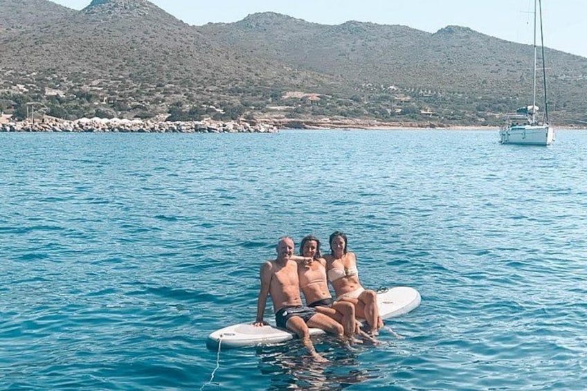 Three people on a watercraft in a body of water with a sailboat in the background and a mountainous landscape. 5