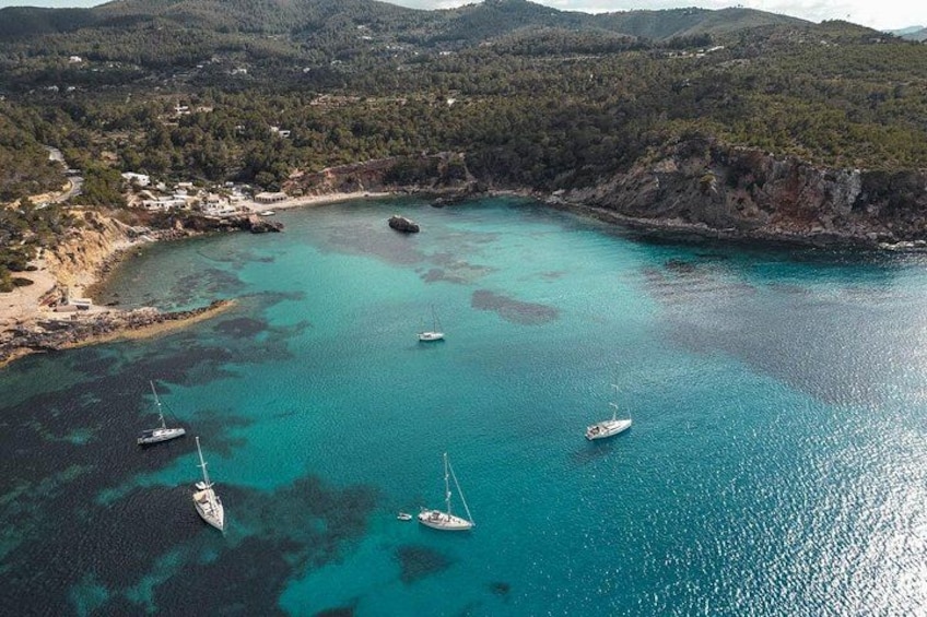 A turquoise bay with boats, a rocky coastline, and a forested hill in the background. 3