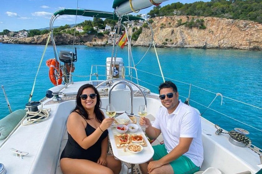 A couple on a sailboat with a plate of food and drinks, surrounded by calm waters and a rocky coastline. 1