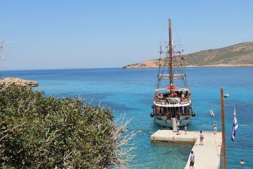 A sailing ship docked at a pier with clear blue water and a hilly landscape in the background. 2