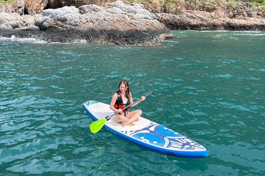 Una persona sentada en una tabla de remo en el agua, con rocas al fondo. 4
