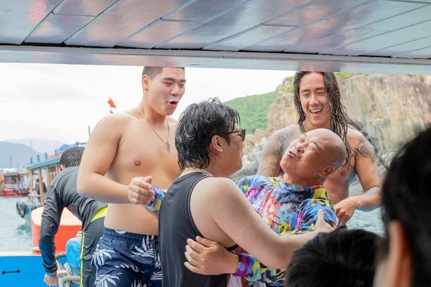 Un grupo de personas en un bote, algunas con traje de baño, con una costa rocosa y botes al fondo. 5