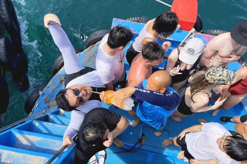 A group of people on a blue boat, some wearing life jackets, others in casual clothing, with a calm sea in the background. 5