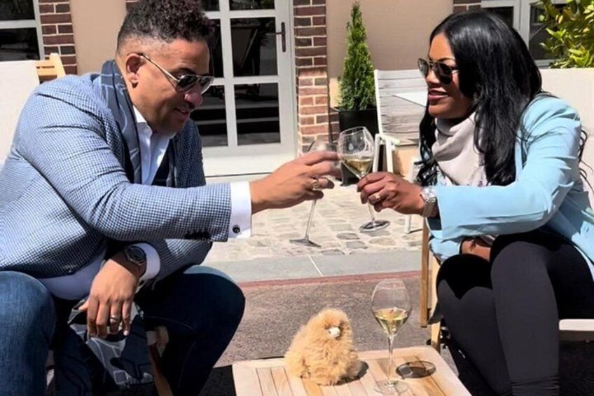 Two people sitting at an outdoor table with a small dog, holding wine glasses. 2