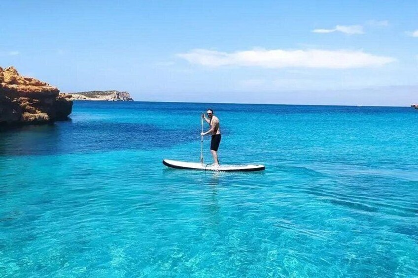 Iemand staat op een paddleboard op helder turquoise water, met rotsachtige kliffen op de achtergrond. 3