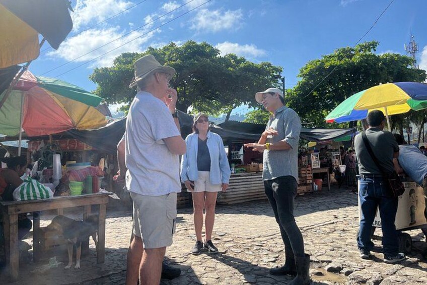 A market scene with people standing and interacting, under colorful umbrellas, and various goods displayed on tables. 3