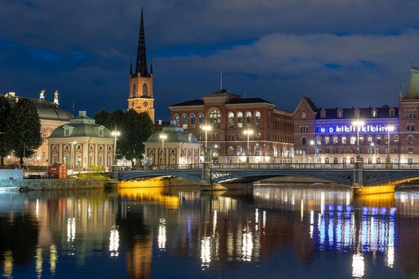 Un paisaje urbano nocturno con una aguja de iglesia destacada, edificios iluminados y un puente sobre un río tranquilo que refleja las luces. 2