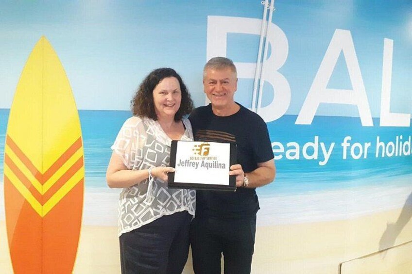Two people holding a plaque in front of a beach-themed backdrop with a surfboard. 1