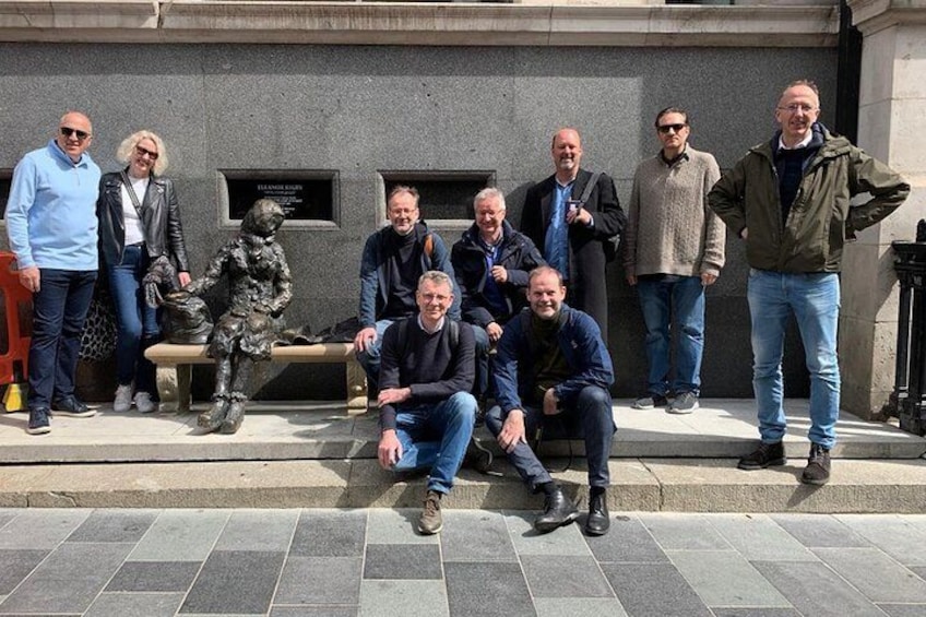 A group of people standing and sitting in front of a statue and a plaque on a stone wall. 2
