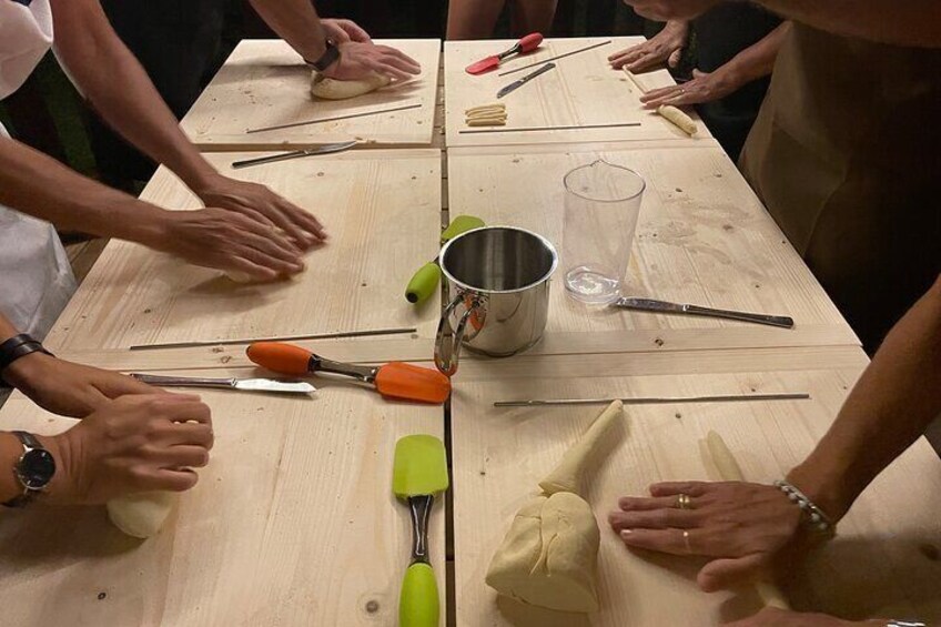 Eine Gruppe von Menschen bereitet Teig zu und verwendet dafür verschiedene Werkzeuge wie Nudelholz und Messer auf einem Holztisch. 5