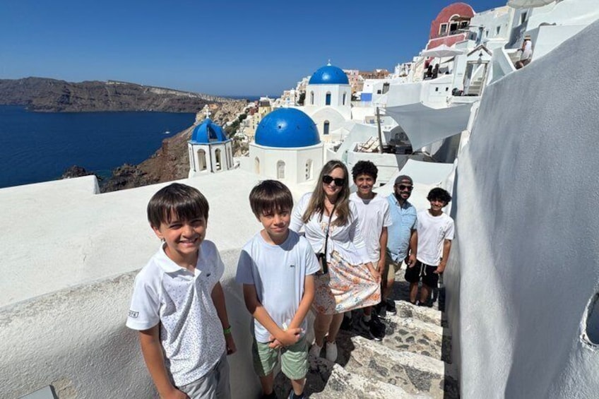 A group of people on a white-washed coastal street with blue domed buildings and a clear blue sea in the background. 5
