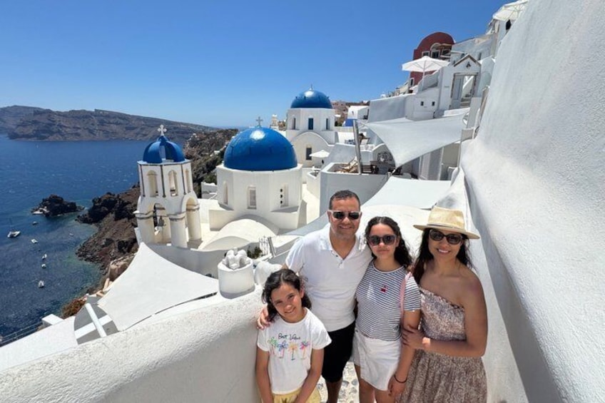 A family on a white-walled street with blue-domed buildings and a clear blue sea in the background. 2
