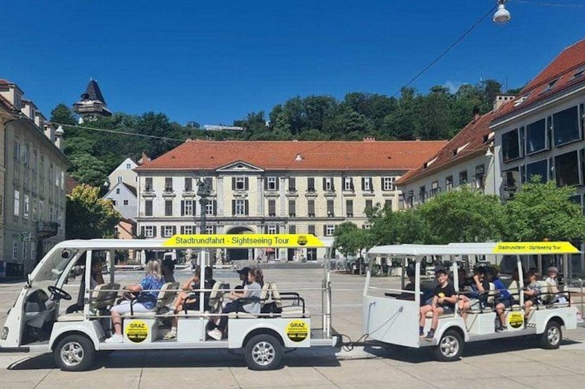 A sightseeing tour vehicle with passengers, parked in a town square with historic buildings in the background. 3