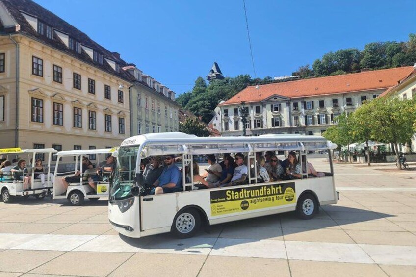Un autobús turístico en una plaza, con pasajeros y edificios históricos al fondo. 4
