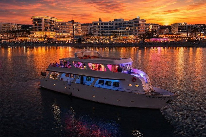A large boat with multiple decks and a lit upper deck, anchored in calm waters with a city skyline in the background. 1
