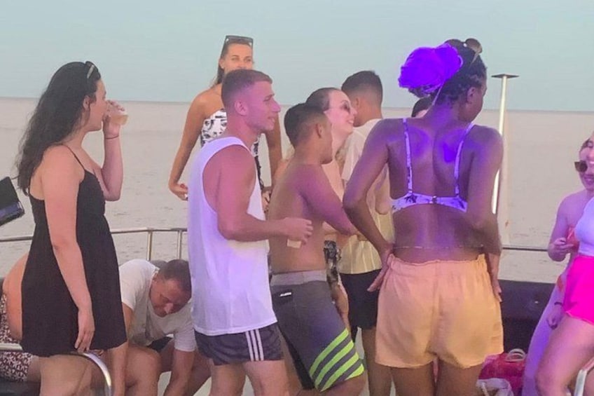 Un gruppo di persone su una barca, alcune in piedi e altre sedute, con un mare calmo sullo sfondo. 2