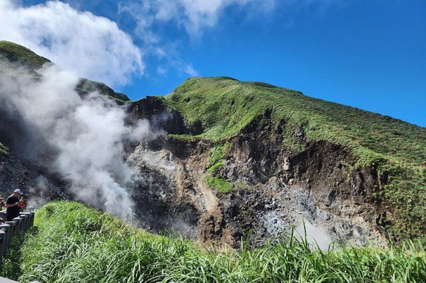 A volcanic landscape with a steam vent, green vegetation, and a clear blue sky. 2