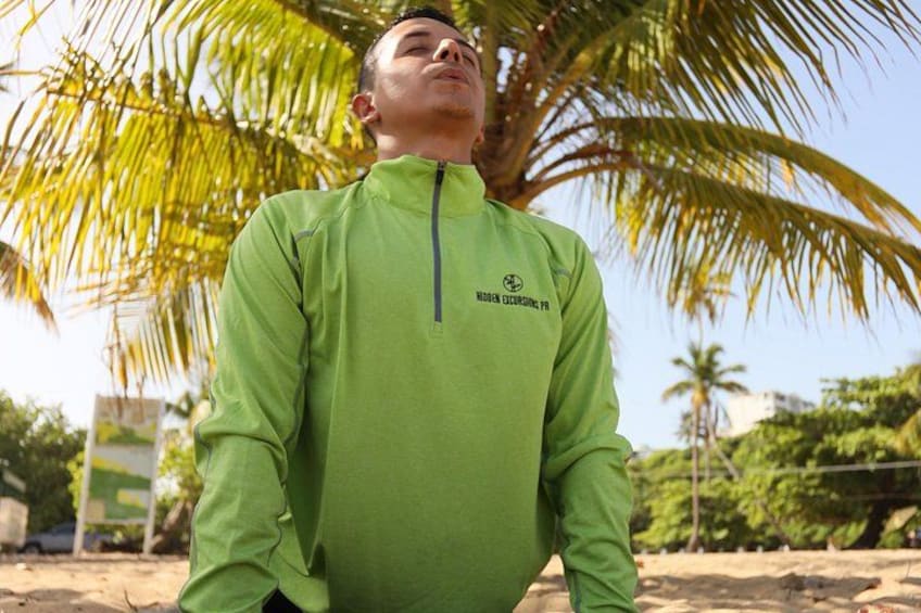 A person wearing a green jacket with a logo, standing outdoors with palm trees in the background. 1