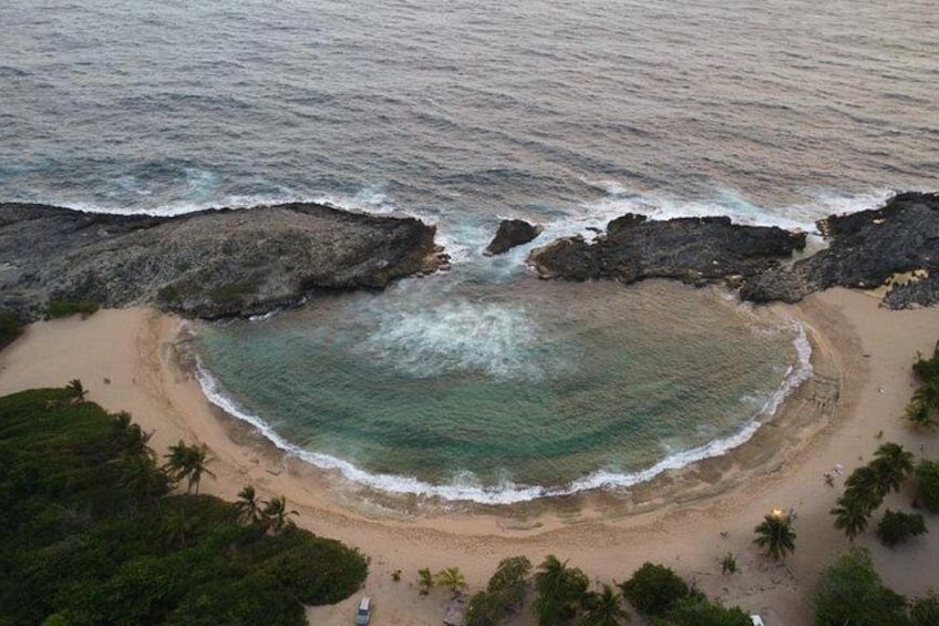A beach with a circular sand formation surrounded by ocean waves, palm trees, and rocky outcrops. 2