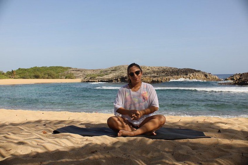 A person sitting cross-legged on a beach with a rocky shoreline and calm sea in the background. 3