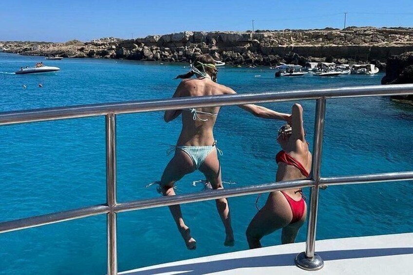 Una persona en traje de baño de pie en la barandilla de un barco, otra persona en traje de baño en el agua, y una costa rocosa con barcos al fondo. 4