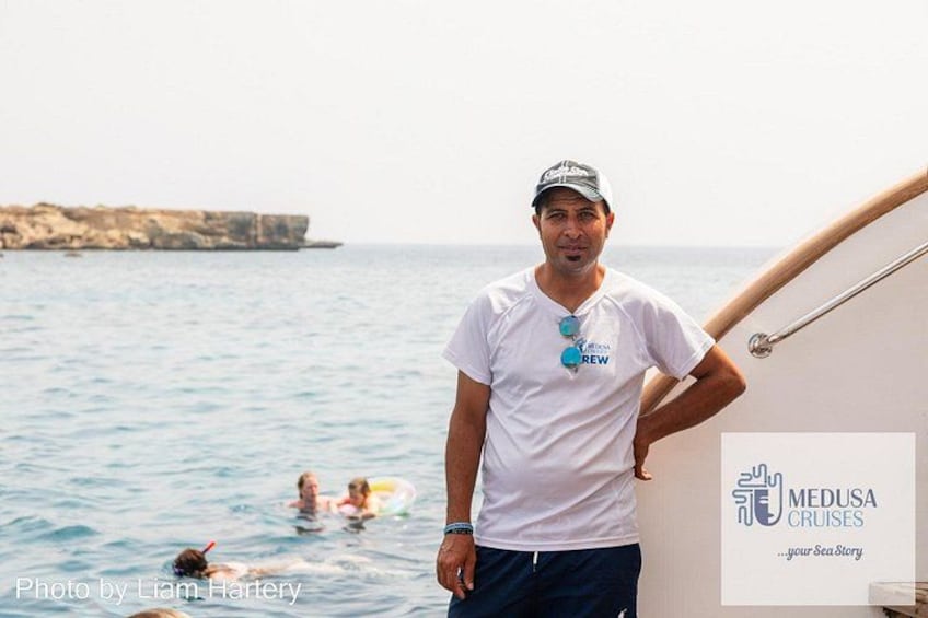 A man standing on a boat with a sea view, a logo for Medusa Cruises, and a rocky coastline in the background. 3