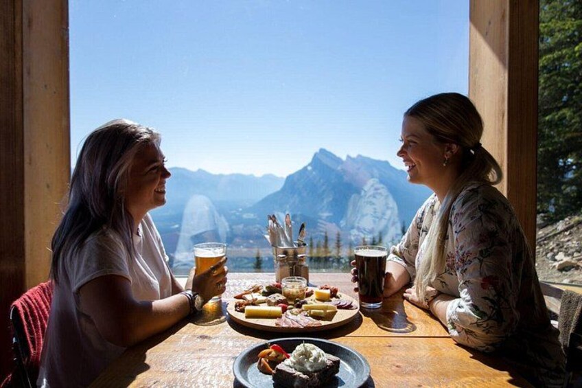 Dos personas sentadas en una mesa de madera con vistas a la montaña, disfrutando de comida y bebidas. 2