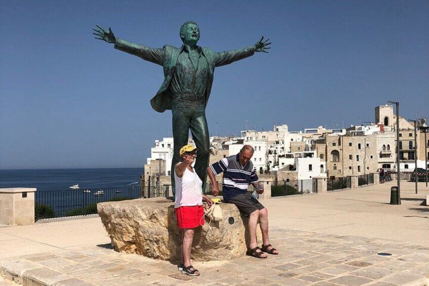 Una estatua de un hombre con los brazos extendidos, una persona de pie a su lado y un paisaje urbano costero al fondo. 3