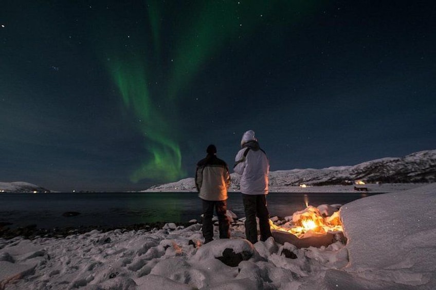 Dos personas junto a una playa nevada con fuego, bajo un cielo nocturno con la aurora boreal. 1