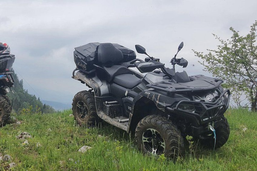 A black ATV with a large storage box on the back, parked on a grassy field with a cloudy sky in the background. 2