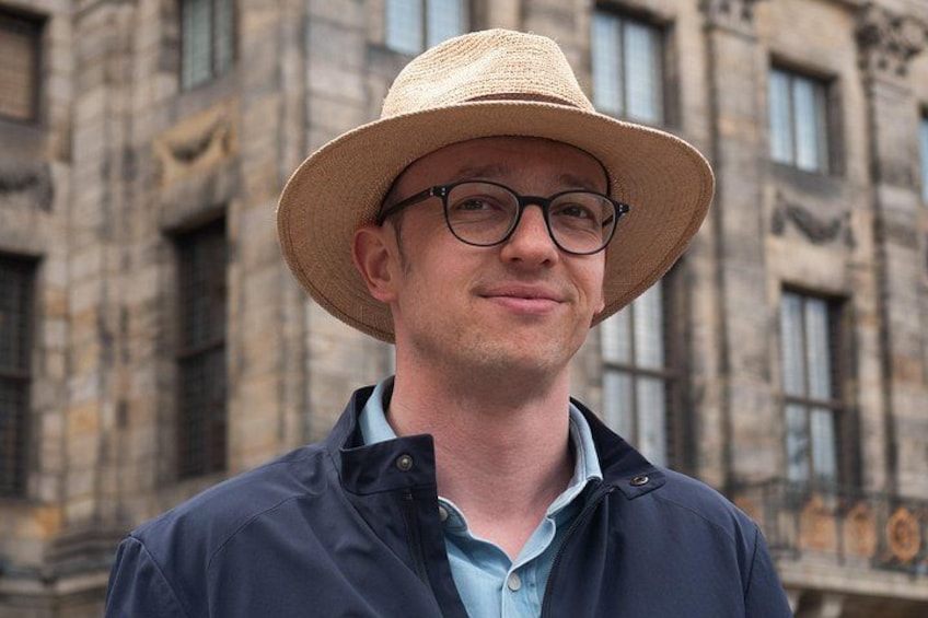En person med en halmhatt och en mörk jacka står framför en byggnad med stora fönster och fasad av sten. 1