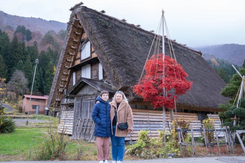 Une maison traditionnelle en bois, avec un toit de chaume très incliné, un arbre aux feuilles rouges et un paysage montagneux en arrière-plan. 3