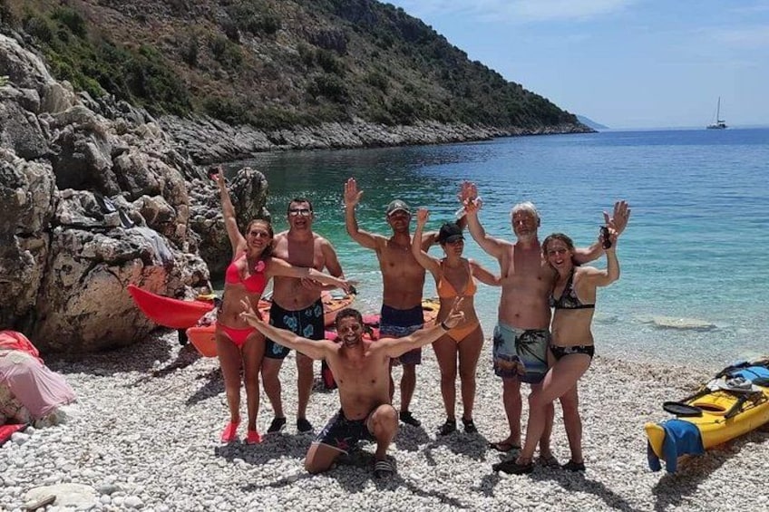 Eine Gruppe Menschen an einem felsigen Strand mit klarem, blauem Wasser und einem Segelboot in der Ferne. 3