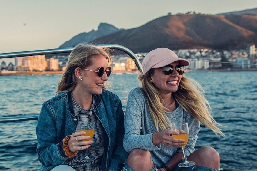 Two people sitting on a boat, holding drinks, with a cityscape and mountains in the background. 2