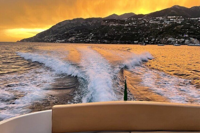 A boat moving through water with waves, a sunset, and a mountainous coastline. 1