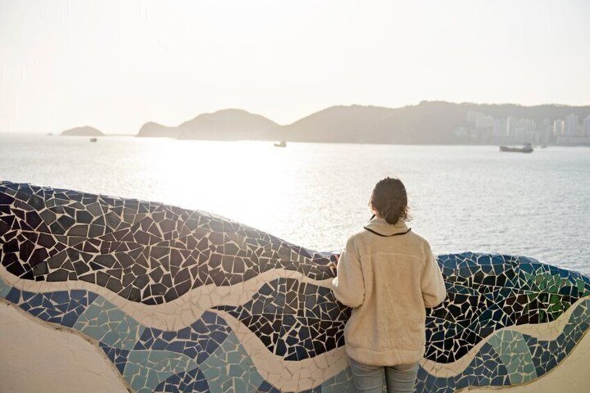 Un mur décoré de mosaïques, une personne debout et une étendue d’eau avec, au loin, une terre ferme et un bateau. 3