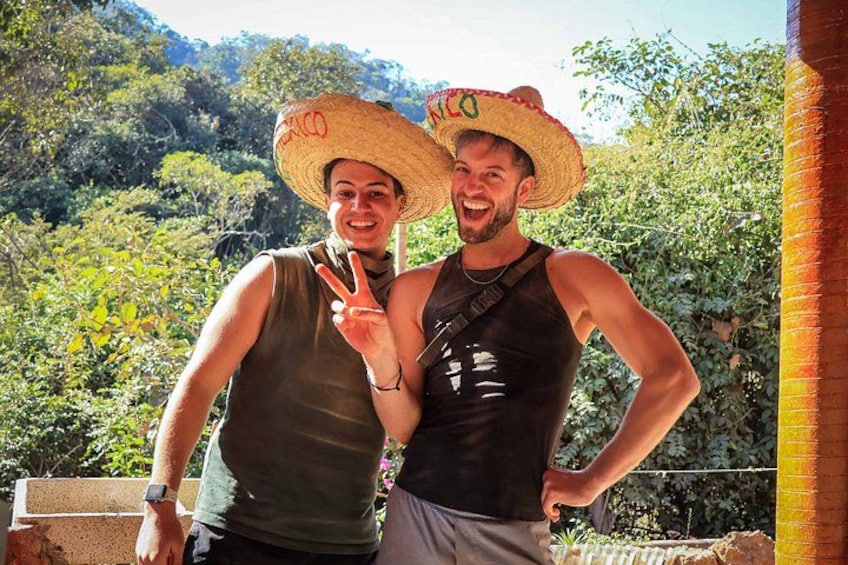 Two people wearing straw hats and tank tops, standing outdoors with greenery and a brick wall in the background. 4