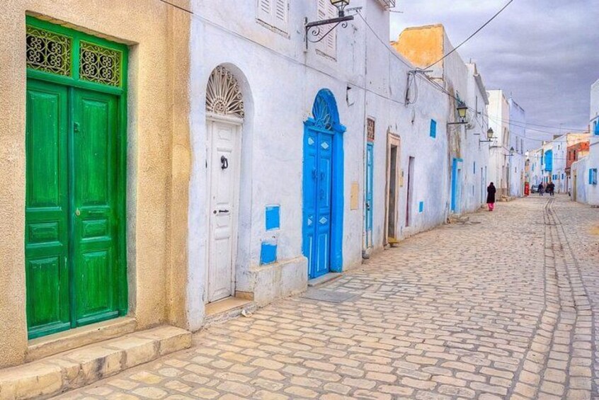 A cobblestone street with white buildings, green and blue doors, and a person walking in the distance. 2
