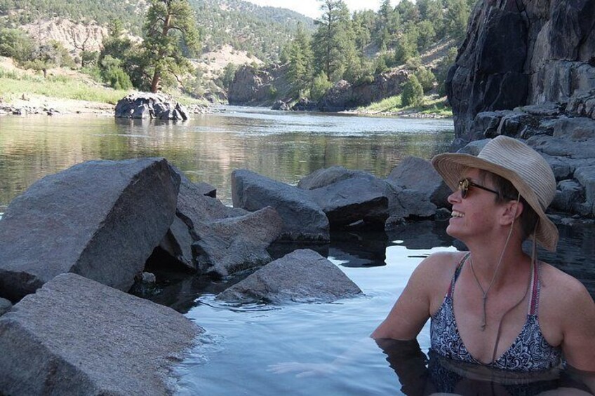 A person sitting by a river with large rocks and a forested hillside in the background. 2