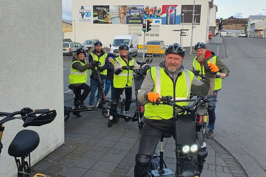 Een groep mensen op elektrische fietsen, met veiligheidsvesten, op een verharde weg met gebouwen en verkeersborden op de achtergrond. 2