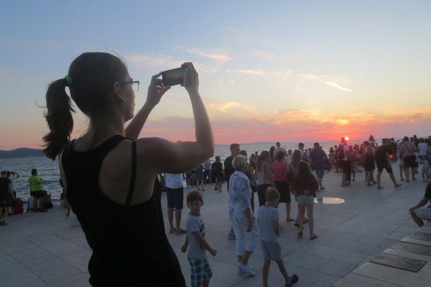 Un coucher de soleil sur une plage bondée, où l’on voit des gens se promener et prendre des photos. 3