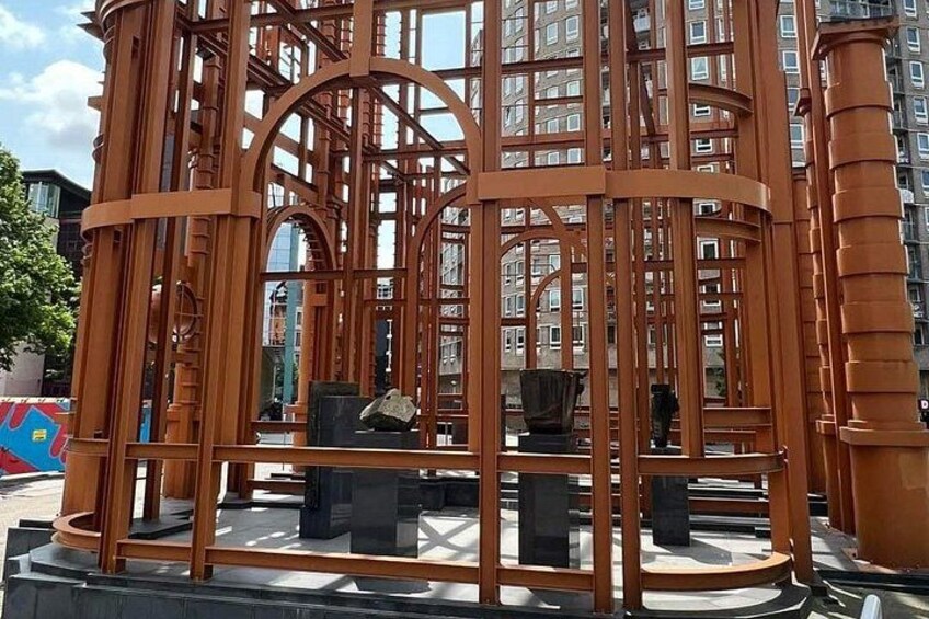 A wooden gazebo with a lattice roof structure, situated in an urban setting with buildings in the background. 5