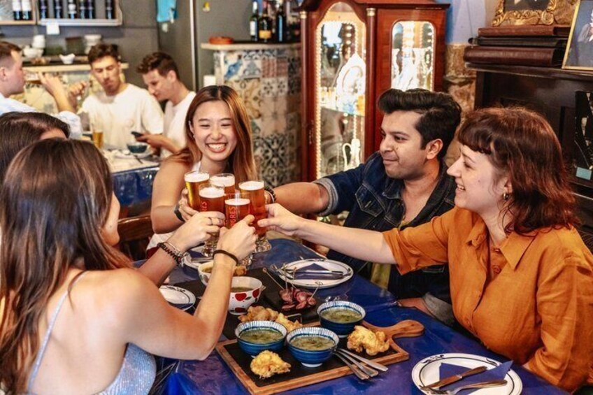 A group of people toasting with beer glasses at a table with bowls of food and a wooden board. 5