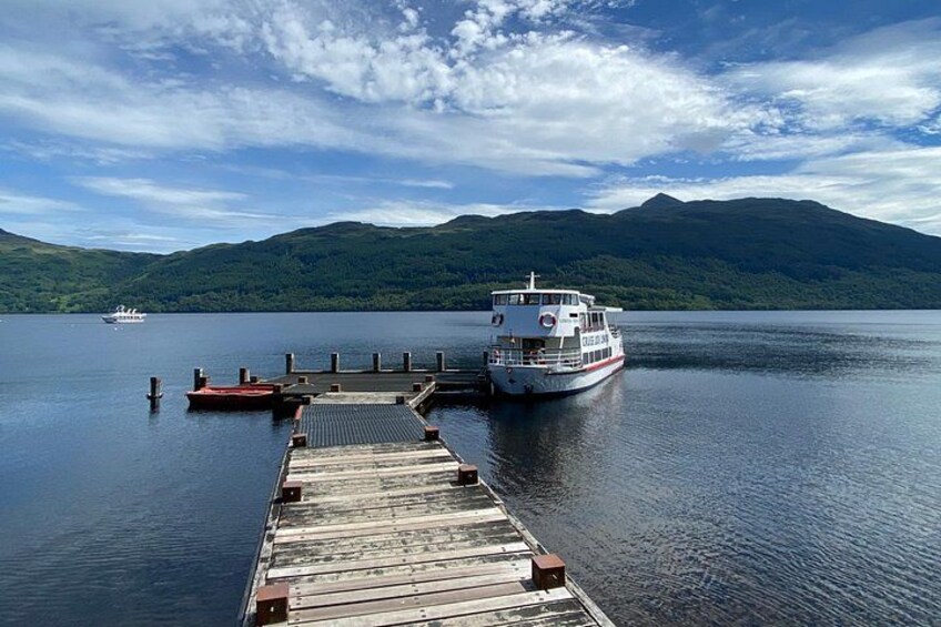 A wooden dock extending into a calm lake with a boat docked at the end. In the background, there are green hills and a partly cloudy sky. 4