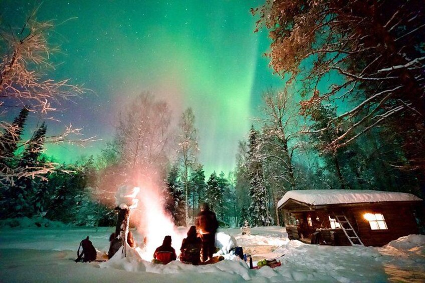 A snowy landscape with a cabin, a fire, and people gathered around. 1