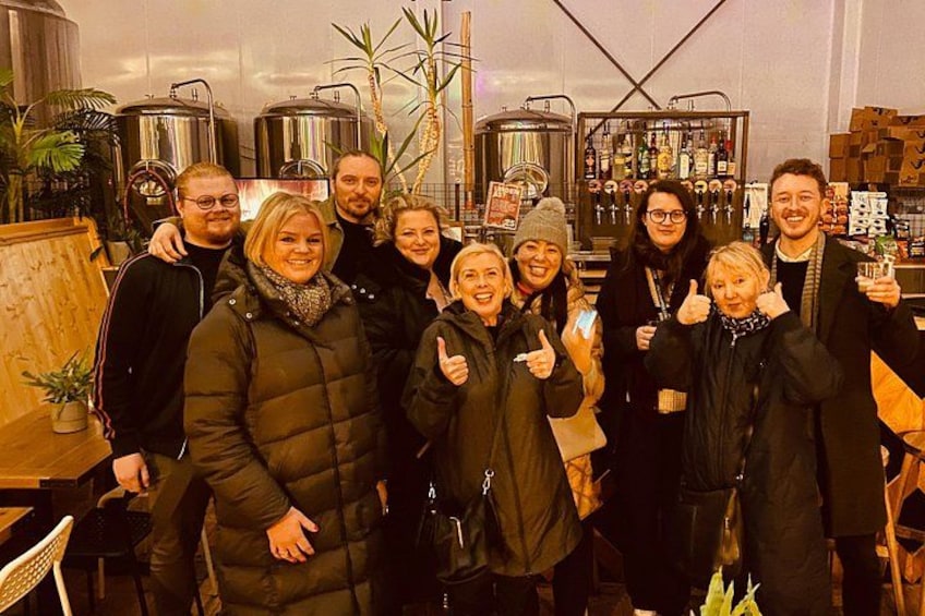 A group of people in a brewery setting with large stainless steel tanks and wooden barrels. 5