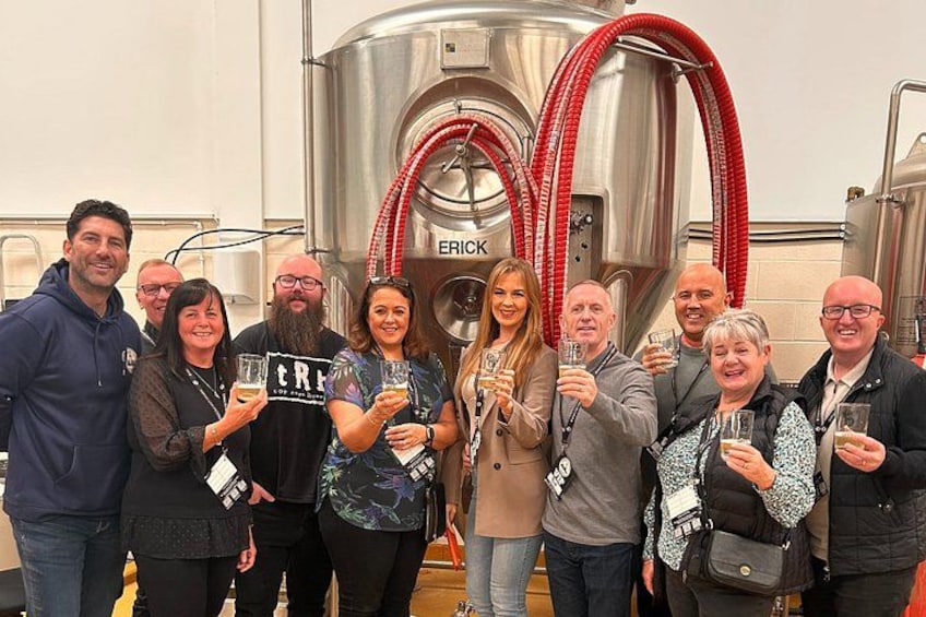 A group of people holding glasses in a brewery setting with stainless steel tanks and red hoses. 4